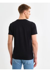 Mladić u jednostavnoj crnoj majici kratkih rukava i svijetloplavim trapericama, okrenut leđima, na bijeloj pozadini.