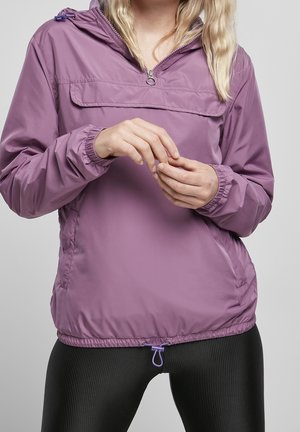 Femme portant une veste violette zippée avec un ourlet ajustable et des leggings noirs texturés, mains se touchant devant, sur un fond gris uni.