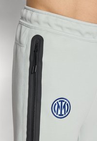 Jasnoszare dresy o tapered fit, z czarnymi bocznymi paskami, kieszenią na zamek i niebieskim haftowanym logo na udzie.
