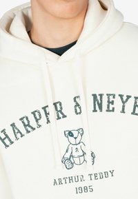 Sudadera cremosa con cordones que presenta el texto impreso "HARPER & NEYER" y una ilustración de un oso de peluche; ajuste relajado, textura suave.