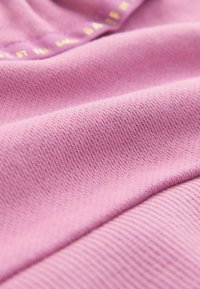Tejido de punto rosa con una superficie texturada, que presenta un patrón acanalado. Textura suave y lisa con un logo en relieve en dorado en el borde.