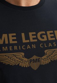Musta puuvillapaita, jossa on kultaisella painetut tekstit "PME LEGEND AMERICAN CLASSIC" ja siipisymboli. Sujuva rakenne, pyöreä kaula-aukko.
