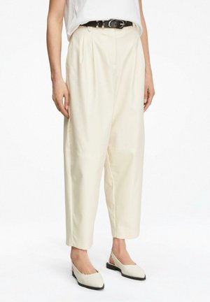 Kobieta ubrana w luźne kremowe spodnie, czarny pasek, biały bezrękawnik oraz kremowe sandały na niskim czarnym obcasie.