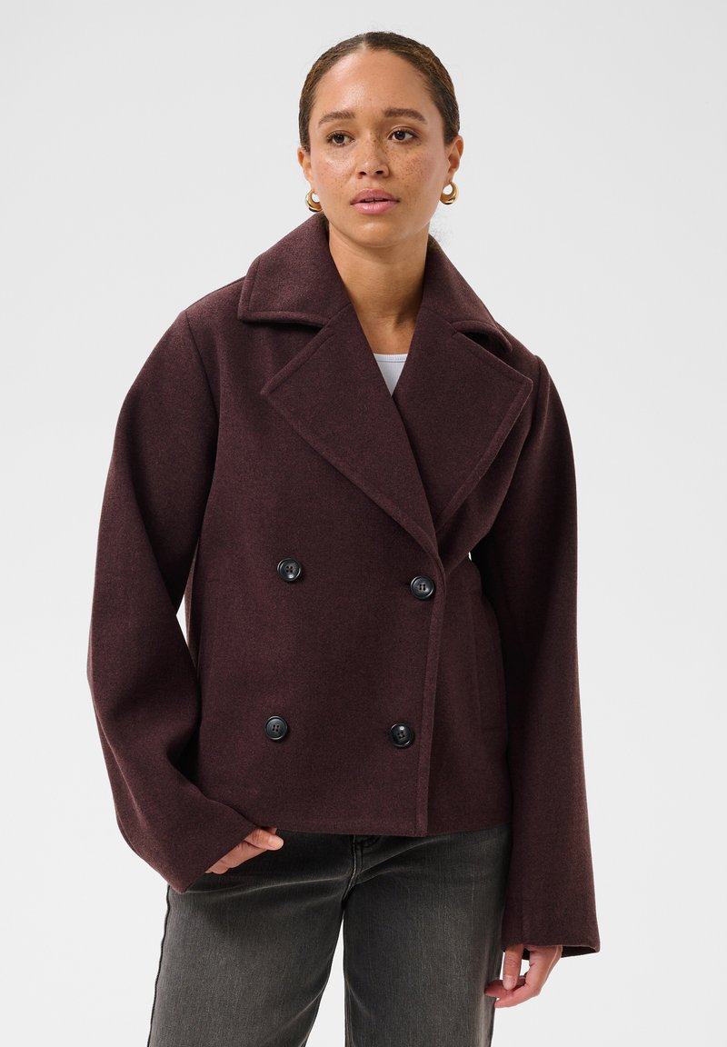 Femme portant un manteau en laine violet foncé à double boutonnage avec de larges revers, boutons noirs, associé à un jean gris foncé et des boucles d'oreilles anneaux dorés.