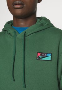 Zielona bluza z kapturem i sznurkiem, z małą, wielokolorową naszywką z logo Nike. Pod spodem znajduje się niebieska koszulka, widoczna przy kołnierzu.
