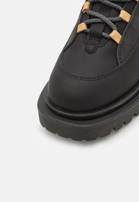 Scarpa da trekking nera in pelle con superficie texture, suola in gomma spessa e lacci grigi accentuati da portaricci arancioni. Design a punta tonda.