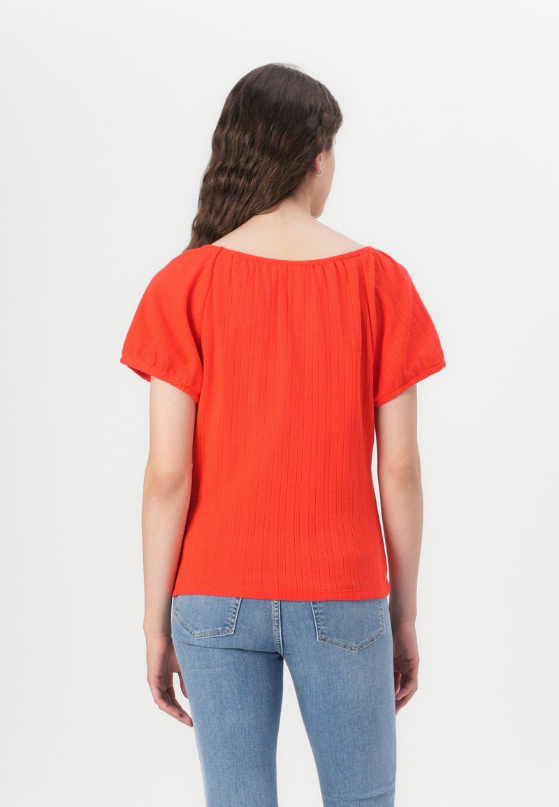 Rotes, kurzärmliges Oberteil mit lockerer Passform, ausgestattet mit einem strukturierten Muster und weitem Ausschnitt, kombiniert mit hellblauen Jeans.