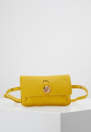 Żółta teksturowana skórzana torba na pasku z klapką i okrągłym złotym zapięciem na guzik, regulowany pasek z metalowymi oczkami, umieszczona na białym piedestale.