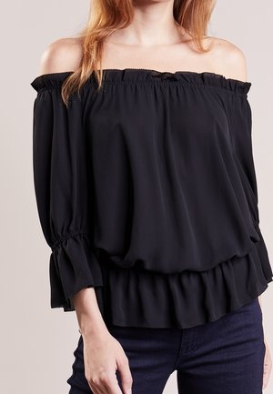 Femme portant un chemisier noir épaules dénudées avec poignets et ourlet froncés, associé à un jean bleu foncé, montrée du cou aux hanches.