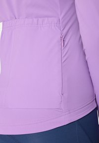 Svijetlolvioletna sportska jakna s džepom na zatvarač i mekim, teksturiranim materijalom. Ima prilagođeni dizajn i krojene šavove za naglašavanje siluete.