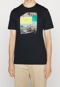 Svart t-shirt med kort ärm med en färgglad grafik av Palm Springs, Kalifornien, bärs av en person med beiga byxor, visad från halsen till mitten av låret.