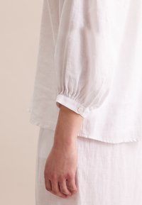 Camicia bianca di lino con maniche lunghe e a sbuffo. Presenta un bottone al polsino, una texture leggera e una vestibilità rilassata, abbinata a un capo simile.