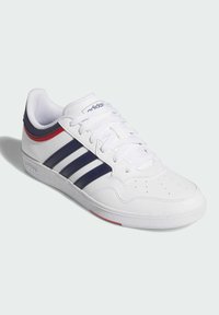 Białe sneakersy z tkaniny i materiału syntetycznego, z granatowymi paskami, czerwonymi akcentami na pięcie oraz teksturowaną gumową podeszwą.