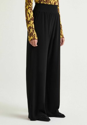 Black wide-leg pants with a smocked waistband, made from smooth fabric. Paired with a long-sleeve patterned top and embellished black shoes.