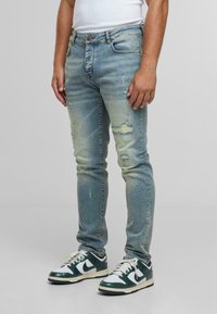 Hombre vistiendo vaqueros desgastados de color azul claro, zapatillas blancas y verdes, y una camiseta sin mangas blanca, de pie sobre una superficie gris.