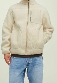 Persona che indossa una giacca in pile color crema con zip anteriore, tasca sul petto con zip e due ampie tasche laterali, abbinata a jeans azzurri chiari.