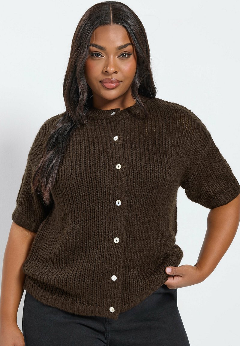 Frau mit langen dunklen Haaren, die eine kurzärmelige, dunkelbraune Strickjacke mit weißen Knöpfen trägt und mit einer Hand in der Hüfte posiert.