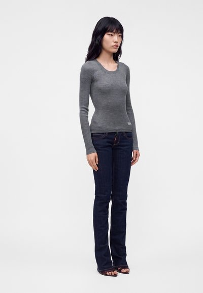 Jeune femme debout portant un pull gris ajusté à manches longues, un jean bleu foncé et des talons ouverts sur un fond clair uni.