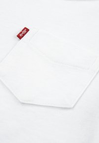 Biały bawełniany materiał z wyraźną kieszenią na piersi i małą czerwoną metką przyszytą nieco poniżej górnej krawędzi. Tekstura jest gładka i miękka.