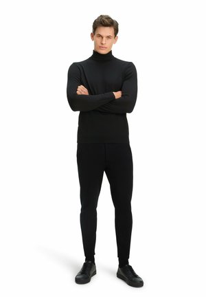 Outfit con dolcevita nero e pantaloni aderenti, realizzato in tessuto morbido. Modello indossa sneakers nere, con le braccia incrociate, su uno sfondo bianco.