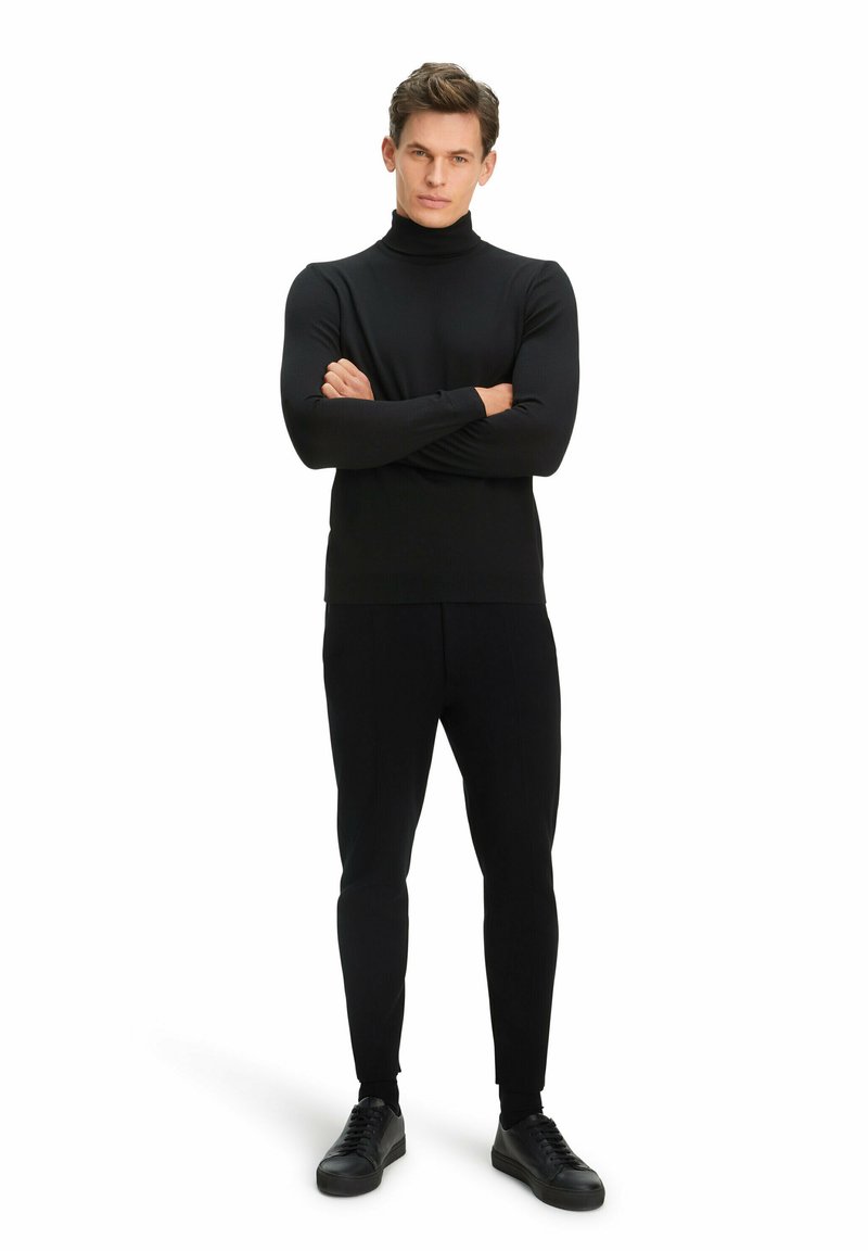 Schwarzer Rollkragenpullover und enge Hose, aus glattem Stoff. Das Model trägt schwarze Sneaker, die Arme verschränkt, vor einem weißen Hintergrund.