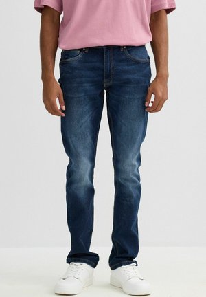 Vaqueros de denim azul oscuro con un corte Slim, ligeramente desgastados en la parte frontal y un diseño clásico de cinco bolsillos. Combinados con zapatillas blancas y una camisa rosa.