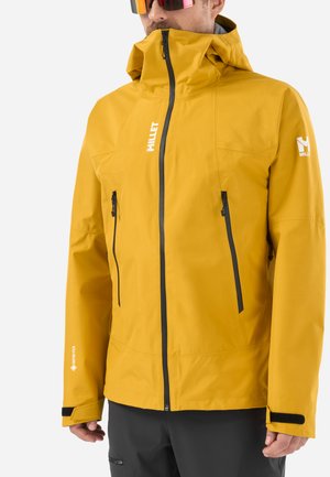 Homme portant une veste imperméable jaune moutarde avec capuche, poches zippées noires et pantalon noir, montré de la moitié du torse à la tête.