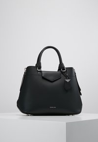 Sac à main en cuir noir avec poignées tressées, poche avant à rabat et étiquette de marque, reposant sur un socle blanc sur fond uni.
