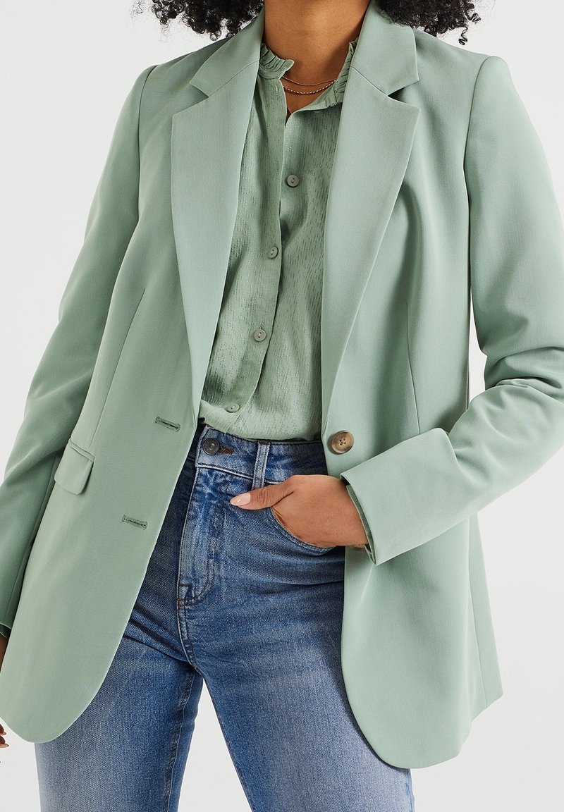 Femme portant un blazer vert clair sur une blouse verte à motifs rentrée dans un jean bleu taille haute, main dans la poche, sur fond blanc.