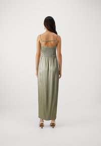 Robe maxi en satin vert olive avec des bretelles fines, un dos smocké et une silhouette fluide. Portée avec des talons blancs à lanières.