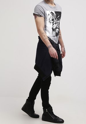 Graues Baumwoll-T-Shirt mit Grafikdruck, schwarzer liegender Sweatshirt um die Taille gebunden, schwarze Slim-Fit-Jeans und schwarze High-Top-Sneaker.