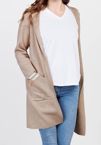 Kvinna klädd i en beige öppen cardigan med framficka, vit V-ringad T-shirt och blå jeans mot en enfärgad bakgrund.