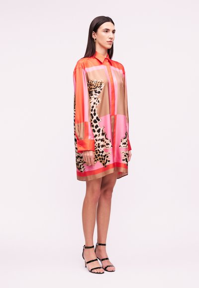 Γυναίκα που φοράει μακρυμάνικο, φαρδύ φόρεμα πουκαμίσα με πορτοκαλί, ροζ, καφέ και λεοπάρ σχέδια, συνδυασμένο με μαύρα σανδάλια με τακούνι.