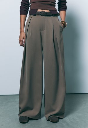 Pantalons larges en tissu taupe avec des plis avant et des poches latérales ; associés à une ceinture sombre et des chaussures simples.