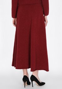 Femeie îmbrăcată cu o fustă lungă din lână bordeaux și pantofi cu toc din suede negru, stând pe un fundal alb.