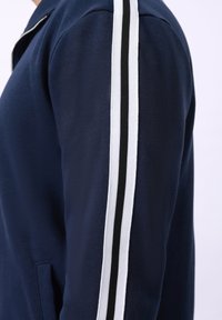 Veste zippée marine en tissu texturé, avec des rayures blanches et noires sur les côtés, et une poche à l'avant du côté gauche.