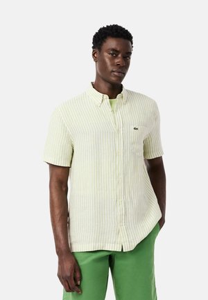 Hombre con camisa de manga corta, botones y rayas amarillas pálidas y pantalones verde brillante, de pie con una mano en el bolsillo sobre un fondo liso.