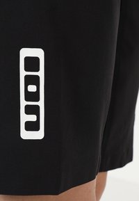 Mustikka shortsit, jotka on valmistettu sileästä kankaasta. Niissä on suuret valkoiset logot suunnittelut vasemmalla puolella, joissa on geometrisia muotoja ja aukkoja.