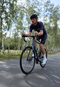 Cestná bicykel so sivým rámom, diskovými brzdami a tenkými čiernymi pneumatikami; cyklista v tmavom dresu a šortkách jazdí po asfaltovej ceste medzi stromami.