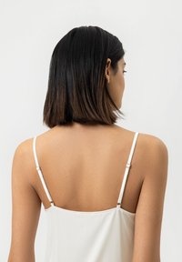 Débardeur blanc avec des bretelles spaghetti ajustables et des garnitures dorées, présentant une texture lisse et une coupe ajustée.