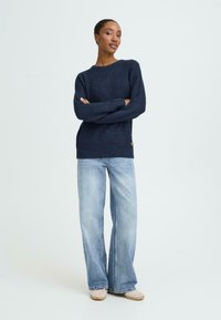Marineblå strikket trøje med rund halsudskæring, ribbet tekstur og en lille logo-detalje, parret med lyseblå flared jeans.