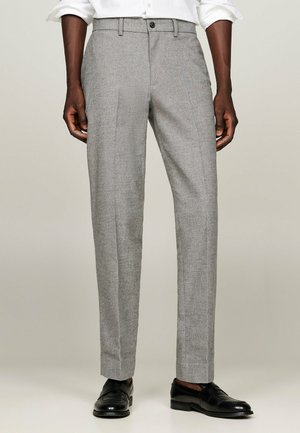 Pantalon gris ajusté en tissu texturé, avec une coupe droite, des plis sur le devant et une fermeture à un bouton. Des chaussures en cuir noir complètent le look.
