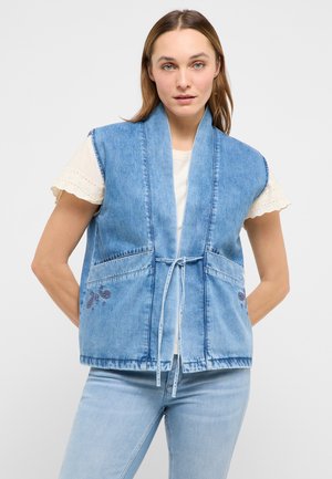 Frau trägt eine ärmellose, hellblaue Jeansweste, die in der Taille gebunden ist, über einem weißen Kurzarm-Top mit Spitzenbesatz, kombiniert mit hellblauen Jeans.