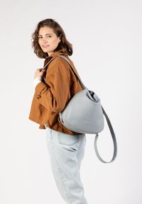 Grijze leren rugzak met een unieke driehoekige vorm, een enkele verstelbare schouderband en een gladde textuur. Het model draagt een bruine jas en lichte jeans.