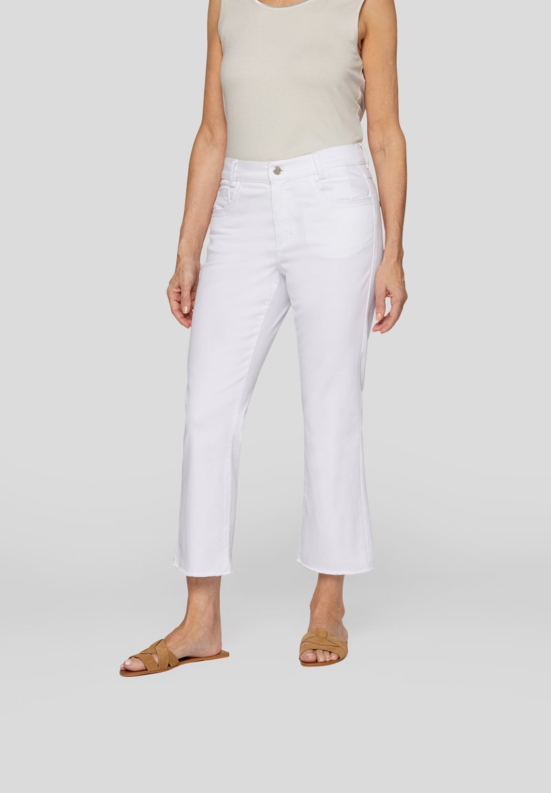 Weiße, hoch geschnittene Jeans mit einer enganliegenden Taille und fransigem Saum, hergestellt aus Denim-Stoff. Getragen mit einem beigen Tanktop und braunen Slippern.