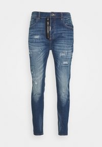 Vaqueros de mezclilla azul oscuro con corte slim, que presentan parches desgastados, salpicaduras de pintura blanca y un detalle de bolsillo con cremallera en la parte delantera.