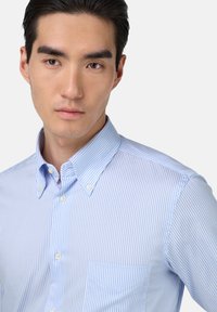 Jasnoniebieska koszula z guzikami, w cienkie pionowe białe paski, z klasycznym kołnierzykiem i małą kieszonką na piersi.