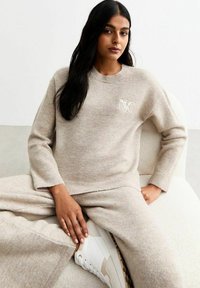 Pull en tricot beige à col rond, arborant un logo brodé "NYC". Associé à une jupe en tricot assortie et des baskets blanches.