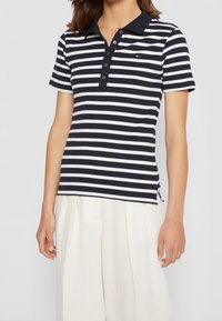 Marineblauw en wit gestreepte polo shirt met korte mouwen, een kraag en een sluiting met drie knopen; beschikt over het merkembleem op de borst en zijkleppen.