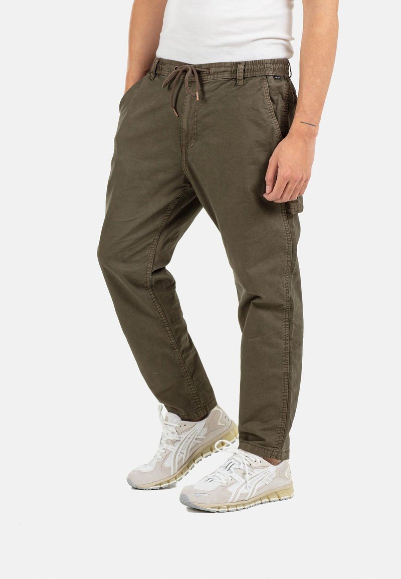 Man wearing olive green casual trousers with drawstring waist and white sneakers, standing against a plain white background.